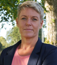 Jennie Cederholm Björklund har mörk kavaj och rosa topp. Hon står ute i naturen med ett träd vid sidan. 