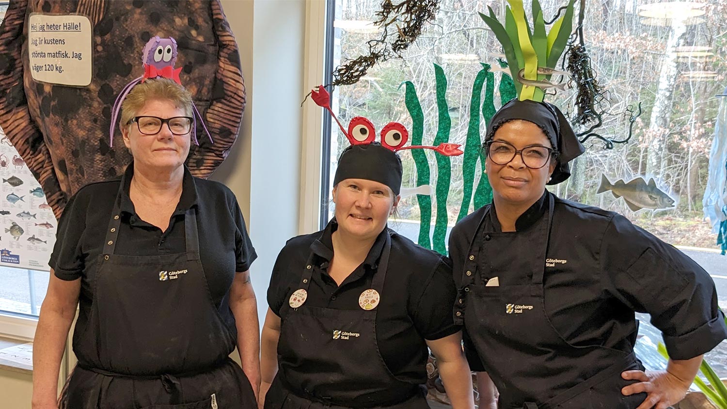 Tre glada kockar poserar i huvudbonader med havstema, framför en dekorativ undervattensscen.