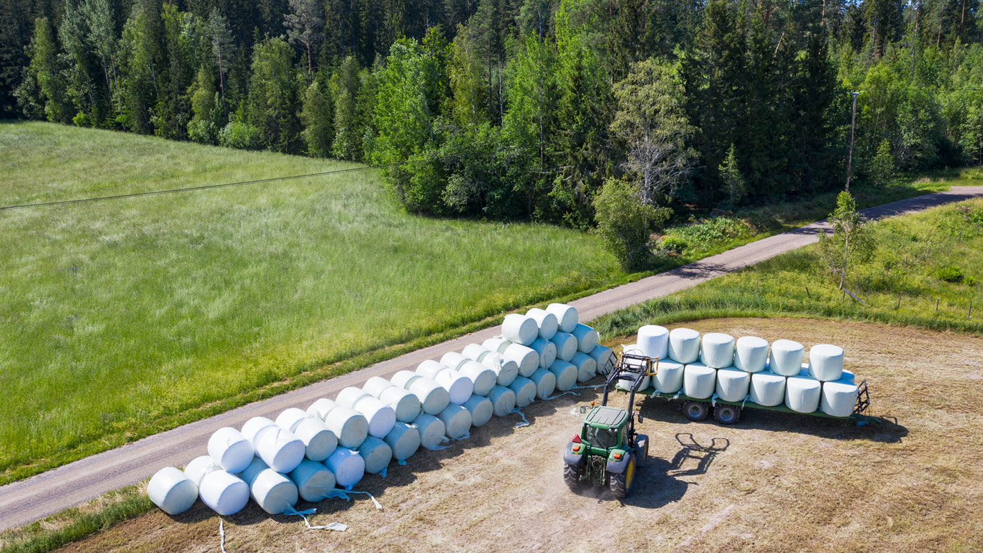 Vy över ett fält där en traktor lastar ensilagebalar