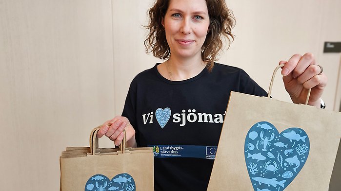Kvinna håller i papperskassar med blått hjärta, på t-shirten står texten Vi "hjärta" sjömat.