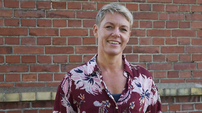 Jennie Cederholm Björklund har en blommig skjorta på sig och står framför en tegelbyggnad