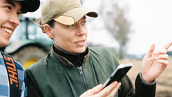 En kvinna i gröna arbetskläder står på en åker och har en mobiltelefon i handen. 