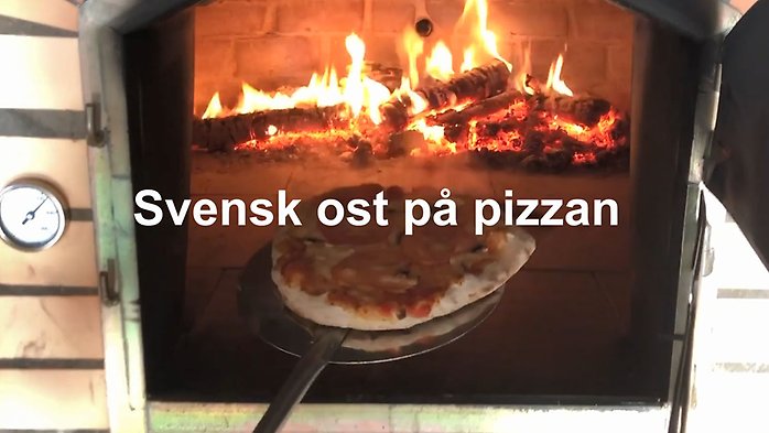 Elden brinner i vedeldad pizzaung. Någon tar ut en pizza ur ugnen med hjälp av en spatel.
