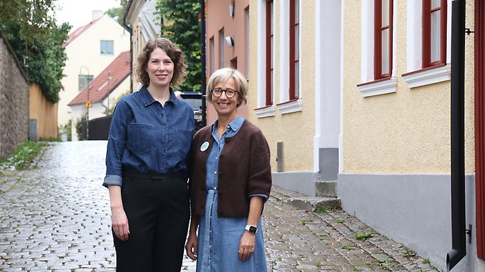 Två kvinnor ler mot kameran på liten gata utomhus.