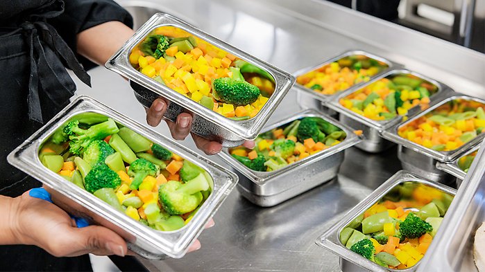 Kock håller i små kantiner med broccoli och andra grönsaker.